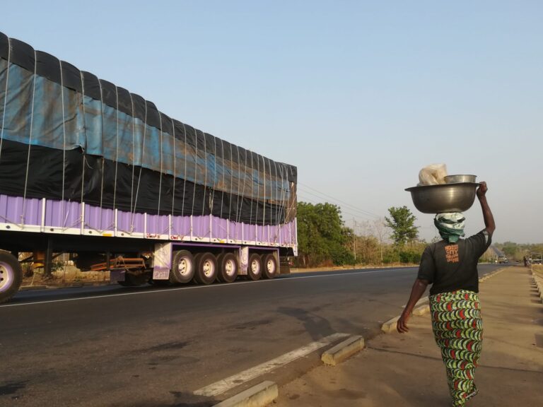 Straße in Westafrika mit traditionell gekleideter Frau mit Korb auf Kopf und vorbefahrendem Lkw